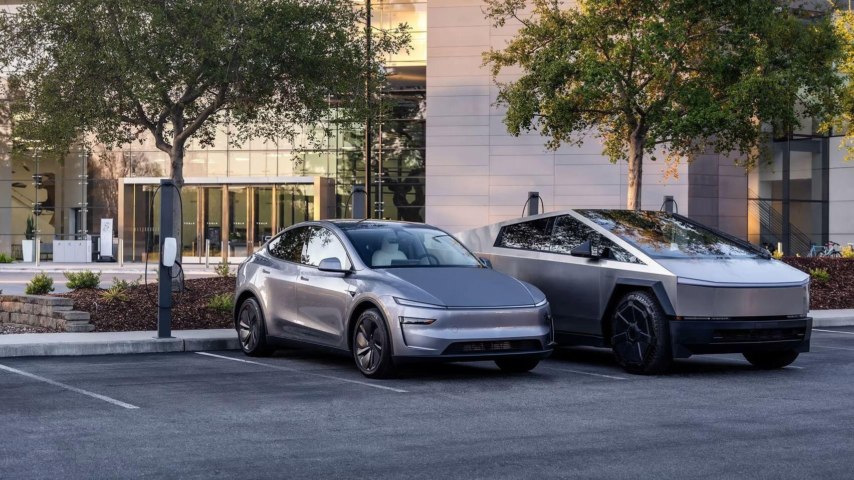 Tesla SUV and Cybertruck parked at chargers in front of a glass office building